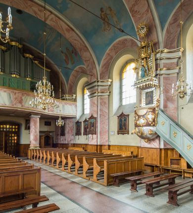 Zdjęcie nr 8: Murowany kościół otrzymał układ jednonawowej świątyni z węższym prezbiterium zwróconym ku północy i wieżą dostawioną do nawy od południa. Prezbiterium jest jednoprzęsłowe zakończone spłaszczoną apsydą. Na jego przedłużeniu usytuowano czworoboczną zakrystię. Prostokątna nawa jest trójprzęsłowa o zaokrąglonych narożnikach. Od frontu wzniesiono czworoboczną wieżę o ściętych narożnikach, flankowaną piętrowymi lokalnościami. Wysunięto ją lekko ku przodowi, gdzie przechodzi wklęsłymi płaszczyznami w boczne części fasady. Jej górne piętro nakryte cebulastym hełmem zostało opięte po bokach wolutami, obok których ustawiono kamienne klasycystyczne wazy. Elewacje zewnętrzne artykułowano podziałami ramowymi, zamykając wysokości ścian gzymsem gurtowy. Na końcu nawy na siodłowym dachu pokrytym blachą umieszczono czworoboczną wieżyczkę na sygnaturkę zwieńczoną cebulastym hełmem z krzyżem. Podobnym dachem nakryto prezbiterium. Wnętrze kościoła zostało rozczłonkowane lizenami z wklęsłymi półlizenami po bokach, ponad którymi rozciągnięto odcinki belkowania. Całość nakryto sklepieniem żaglastym na zdwojonych gurtach. Od południa w nawie urządzono chór muzyczny wsparty na dwóch kolumnach z parapetem wklęsłowypukłym. W prezbiterium usytuowano ołtarz główny, a w nawie dwa ołtarze boczne. Tu także na ścianie zachodniej zawieszono ambonę.  