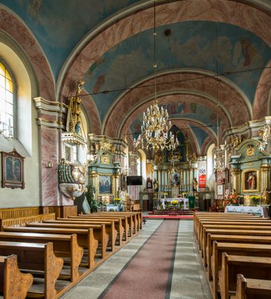 Zdjęcie nr 11: Murowany kościół otrzymał układ jednonawowej świątyni z węższym prezbiterium zwróconym ku północy i wieżą dostawioną do nawy od południa. Prezbiterium jest jednoprzęsłowe zakończone spłaszczoną apsydą. Na jego przedłużeniu usytuowano czworoboczną zakrystię. Prostokątna nawa jest trójprzęsłowa o zaokrąglonych narożnikach. Od frontu wzniesiono czworoboczną wieżę o ściętych narożnikach, flankowaną piętrowymi lokalnościami. Wysunięto ją lekko ku przodowi, gdzie przechodzi wklęsłymi płaszczyznami w boczne części fasady. Jej górne piętro nakryte cebulastym hełmem zostało opięte po bokach wolutami, obok których ustawiono kamienne klasycystyczne wazy. Elewacje zewnętrzne artykułowano podziałami ramowymi, zamykając wysokości ścian gzymsem gurtowy. Na końcu nawy na siodłowym dachu pokrytym blachą umieszczono czworoboczną wieżyczkę na sygnaturkę zwieńczoną cebulastym hełmem z krzyżem. Podobnym dachem nakryto prezbiterium. Wnętrze kościoła zostało rozczłonkowane lizenami z wklęsłymi półlizenami po bokach, ponad którymi rozciągnięto odcinki belkowania. Całość nakryto sklepieniem żaglastym na zdwojonych gurtach. Od południa w nawie urządzono chór muzyczny wsparty na dwóch kolumnach z parapetem wklęsłowypukłym. W prezbiterium usytuowano ołtarz główny, a w nawie dwa ołtarze boczne. Tu także na ścianie zachodniej zawieszono ambonę.  