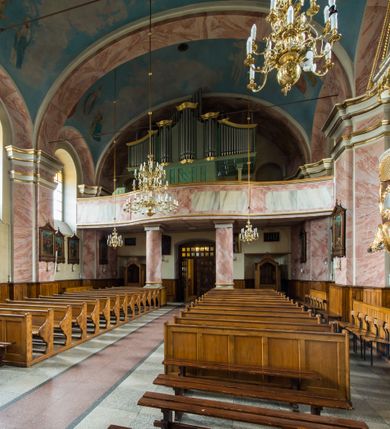 Zdjęcie nr 12: Murowany kościół otrzymał układ jednonawowej świątyni z węższym prezbiterium zwróconym ku północy i wieżą dostawioną do nawy od południa. Prezbiterium jest jednoprzęsłowe zakończone spłaszczoną apsydą. Na jego przedłużeniu usytuowano czworoboczną zakrystię. Prostokątna nawa jest trójprzęsłowa o zaokrąglonych narożnikach. Od frontu wzniesiono czworoboczną wieżę o ściętych narożnikach, flankowaną piętrowymi lokalnościami. Wysunięto ją lekko ku przodowi, gdzie przechodzi wklęsłymi płaszczyznami w boczne części fasady. Jej górne piętro nakryte cebulastym hełmem zostało opięte po bokach wolutami, obok których ustawiono kamienne klasycystyczne wazy. Elewacje zewnętrzne artykułowano podziałami ramowymi, zamykając wysokości ścian gzymsem gurtowy. Na końcu nawy na siodłowym dachu pokrytym blachą umieszczono czworoboczną wieżyczkę na sygnaturkę zwieńczoną cebulastym hełmem z krzyżem. Podobnym dachem nakryto prezbiterium. Wnętrze kościoła zostało rozczłonkowane lizenami z wklęsłymi półlizenami po bokach, ponad którymi rozciągnięto odcinki belkowania. Całość nakryto sklepieniem żaglastym na zdwojonych gurtach. Od południa w nawie urządzono chór muzyczny wsparty na dwóch kolumnach z parapetem wklęsłowypukłym. W prezbiterium usytuowano ołtarz główny, a w nawie dwa ołtarze boczne. Tu także na ścianie zachodniej zawieszono ambonę.  