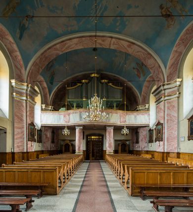 Zdjęcie nr 9: Murowany kościół otrzymał układ jednonawowej świątyni z węższym prezbiterium zwróconym ku północy i wieżą dostawioną do nawy od południa. Prezbiterium jest jednoprzęsłowe zakończone spłaszczoną apsydą. Na jego przedłużeniu usytuowano czworoboczną zakrystię. Prostokątna nawa jest trójprzęsłowa o zaokrąglonych narożnikach. Od frontu wzniesiono czworoboczną wieżę o ściętych narożnikach, flankowaną piętrowymi lokalnościami. Wysunięto ją lekko ku przodowi, gdzie przechodzi wklęsłymi płaszczyznami w boczne części fasady. Jej górne piętro nakryte cebulastym hełmem zostało opięte po bokach wolutami, obok których ustawiono kamienne klasycystyczne wazy. Elewacje zewnętrzne artykułowano podziałami ramowymi, zamykając wysokości ścian gzymsem gurtowy. Na końcu nawy na siodłowym dachu pokrytym blachą umieszczono czworoboczną wieżyczkę na sygnaturkę zwieńczoną cebulastym hełmem z krzyżem. Podobnym dachem nakryto prezbiterium. Wnętrze kościoła zostało rozczłonkowane lizenami z wklęsłymi półlizenami po bokach, ponad którymi rozciągnięto odcinki belkowania. Całość nakryto sklepieniem żaglastym na zdwojonych gurtach. Od południa w nawie urządzono chór muzyczny wsparty na dwóch kolumnach z parapetem wklęsłowypukłym. W prezbiterium usytuowano ołtarz główny, a w nawie dwa ołtarze boczne. Tu także na ścianie zachodniej zawieszono ambonę.  