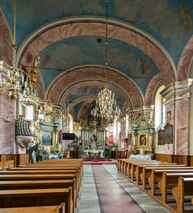 Zdjęcie nr 14: Murowany kościół otrzymał układ jednonawowej świątyni z węższym prezbiterium zwróconym ku północy i wieżą dostawioną do nawy od południa. Prezbiterium jest jednoprzęsłowe zakończone spłaszczoną apsydą. Na jego przedłużeniu usytuowano czworoboczną zakrystię. Prostokątna nawa jest trójprzęsłowa o zaokrąglonych narożnikach. Od frontu wzniesiono czworoboczną wieżę o ściętych narożnikach, flankowaną piętrowymi lokalnościami. Wysunięto ją lekko ku przodowi, gdzie przechodzi wklęsłymi płaszczyznami w boczne części fasady. Jej górne piętro nakryte cebulastym hełmem zostało opięte po bokach wolutami, obok których ustawiono kamienne klasycystyczne wazy. Elewacje zewnętrzne artykułowano podziałami ramowymi, zamykając wysokości ścian gzymsem gurtowy. Na końcu nawy na siodłowym dachu pokrytym blachą umieszczono czworoboczną wieżyczkę na sygnaturkę zwieńczoną cebulastym hełmem z krzyżem. Podobnym dachem nakryto prezbiterium. Wnętrze kościoła zostało rozczłonkowane lizenami z wklęsłymi półlizenami po bokach, ponad którymi rozciągnięto odcinki belkowania. Całość nakryto sklepieniem żaglastym na zdwojonych gurtach. Od południa w nawie urządzono chór muzyczny wsparty na dwóch kolumnach z parapetem wklęsłowypukłym. W prezbiterium usytuowano ołtarz główny, a w nawie dwa ołtarze boczne. Tu także na ścianie zachodniej zawieszono ambonę.  