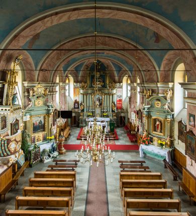 Zdjęcie nr 15: Murowany kościół otrzymał układ jednonawowej świątyni z węższym prezbiterium zwróconym ku północy i wieżą dostawioną do nawy od południa. Prezbiterium jest jednoprzęsłowe zakończone spłaszczoną apsydą. Na jego przedłużeniu usytuowano czworoboczną zakrystię. Prostokątna nawa jest trójprzęsłowa o zaokrąglonych narożnikach. Od frontu wzniesiono czworoboczną wieżę o ściętych narożnikach, flankowaną piętrowymi lokalnościami. Wysunięto ją lekko ku przodowi, gdzie przechodzi wklęsłymi płaszczyznami w boczne części fasady. Jej górne piętro nakryte cebulastym hełmem zostało opięte po bokach wolutami, obok których ustawiono kamienne klasycystyczne wazy. Elewacje zewnętrzne artykułowano podziałami ramowymi, zamykając wysokości ścian gzymsem gurtowy. Na końcu nawy na siodłowym dachu pokrytym blachą umieszczono czworoboczną wieżyczkę na sygnaturkę zwieńczoną cebulastym hełmem z krzyżem. Podobnym dachem nakryto prezbiterium. Wnętrze kościoła zostało rozczłonkowane lizenami z wklęsłymi półlizenami po bokach, ponad którymi rozciągnięto odcinki belkowania. Całość nakryto sklepieniem żaglastym na zdwojonych gurtach. Od południa w nawie urządzono chór muzyczny wsparty na dwóch kolumnach z parapetem wklęsłowypukłym. W prezbiterium usytuowano ołtarz główny, a w nawie dwa ołtarze boczne. Tu także na ścianie zachodniej zawieszono ambonę.  