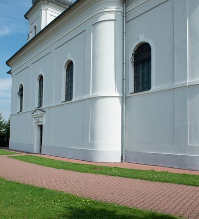 Zdjęcie nr 21: Murowany kościół otrzymał układ jednonawowej świątyni z węższym prezbiterium zwróconym ku północy i wieżą dostawioną do nawy od południa. Prezbiterium jest jednoprzęsłowe zakończone spłaszczoną apsydą. Na jego przedłużeniu usytuowano czworoboczną zakrystię. Prostokątna nawa jest trójprzęsłowa o zaokrąglonych narożnikach. Od frontu wzniesiono czworoboczną wieżę o ściętych narożnikach, flankowaną piętrowymi lokalnościami. Wysunięto ją lekko ku przodowi, gdzie przechodzi wklęsłymi płaszczyznami w boczne części fasady. Jej górne piętro nakryte cebulastym hełmem zostało opięte po bokach wolutami, obok których ustawiono kamienne klasycystyczne wazy. Elewacje zewnętrzne artykułowano podziałami ramowymi, zamykając wysokości ścian gzymsem gurtowy. Na końcu nawy na siodłowym dachu pokrytym blachą umieszczono czworoboczną wieżyczkę na sygnaturkę zwieńczoną cebulastym hełmem z krzyżem. Podobnym dachem nakryto prezbiterium. Wnętrze kościoła zostało rozczłonkowane lizenami z wklęsłymi półlizenami po bokach, ponad którymi rozciągnięto odcinki belkowania. Całość nakryto sklepieniem żaglastym na zdwojonych gurtach. Od południa w nawie urządzono chór muzyczny wsparty na dwóch kolumnach z parapetem wklęsłowypukłym. W prezbiterium usytuowano ołtarz główny, a w nawie dwa ołtarze boczne. Tu także na ścianie zachodniej zawieszono ambonę.  