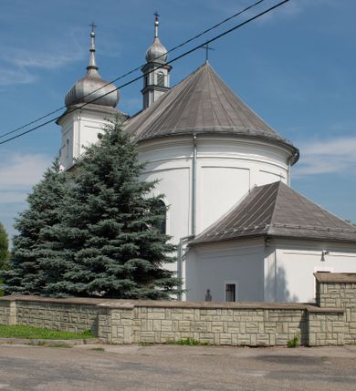 Zdjęcie nr 19: Murowany kościół otrzymał układ jednonawowej świątyni z węższym prezbiterium zwróconym ku północy i wieżą dostawioną do nawy od południa. Prezbiterium jest jednoprzęsłowe zakończone spłaszczoną apsydą. Na jego przedłużeniu usytuowano czworoboczną zakrystię. Prostokątna nawa jest trójprzęsłowa o zaokrąglonych narożnikach. Od frontu wzniesiono czworoboczną wieżę o ściętych narożnikach, flankowaną piętrowymi lokalnościami. Wysunięto ją lekko ku przodowi, gdzie przechodzi wklęsłymi płaszczyznami w boczne części fasady. Jej górne piętro nakryte cebulastym hełmem zostało opięte po bokach wolutami, obok których ustawiono kamienne klasycystyczne wazy. Elewacje zewnętrzne artykułowano podziałami ramowymi, zamykając wysokości ścian gzymsem gurtowy. Na końcu nawy na siodłowym dachu pokrytym blachą umieszczono czworoboczną wieżyczkę na sygnaturkę zwieńczoną cebulastym hełmem z krzyżem. Podobnym dachem nakryto prezbiterium. Wnętrze kościoła zostało rozczłonkowane lizenami z wklęsłymi półlizenami po bokach, ponad którymi rozciągnięto odcinki belkowania. Całość nakryto sklepieniem żaglastym na zdwojonych gurtach. Od południa w nawie urządzono chór muzyczny wsparty na dwóch kolumnach z parapetem wklęsłowypukłym. W prezbiterium usytuowano ołtarz główny, a w nawie dwa ołtarze boczne. Tu także na ścianie zachodniej zawieszono ambonę.  