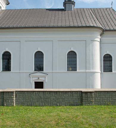 Zdjęcie nr 20: Murowany kościół otrzymał układ jednonawowej świątyni z węższym prezbiterium zwróconym ku północy i wieżą dostawioną do nawy od południa. Prezbiterium jest jednoprzęsłowe zakończone spłaszczoną apsydą. Na jego przedłużeniu usytuowano czworoboczną zakrystię. Prostokątna nawa jest trójprzęsłowa o zaokrąglonych narożnikach. Od frontu wzniesiono czworoboczną wieżę o ściętych narożnikach, flankowaną piętrowymi lokalnościami. Wysunięto ją lekko ku przodowi, gdzie przechodzi wklęsłymi płaszczyznami w boczne części fasady. Jej górne piętro nakryte cebulastym hełmem zostało opięte po bokach wolutami, obok których ustawiono kamienne klasycystyczne wazy. Elewacje zewnętrzne artykułowano podziałami ramowymi, zamykając wysokości ścian gzymsem gurtowy. Na końcu nawy na siodłowym dachu pokrytym blachą umieszczono czworoboczną wieżyczkę na sygnaturkę zwieńczoną cebulastym hełmem z krzyżem. Podobnym dachem nakryto prezbiterium. Wnętrze kościoła zostało rozczłonkowane lizenami z wklęsłymi półlizenami po bokach, ponad którymi rozciągnięto odcinki belkowania. Całość nakryto sklepieniem żaglastym na zdwojonych gurtach. Od południa w nawie urządzono chór muzyczny wsparty na dwóch kolumnach z parapetem wklęsłowypukłym. W prezbiterium usytuowano ołtarz główny, a w nawie dwa ołtarze boczne. Tu także na ścianie zachodniej zawieszono ambonę.  