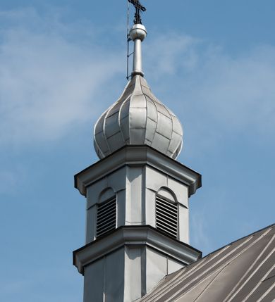 Zdjęcie nr 23: Murowany kościół otrzymał układ jednonawowej świątyni z węższym prezbiterium zwróconym ku północy i wieżą dostawioną do nawy od południa. Prezbiterium jest jednoprzęsłowe zakończone spłaszczoną apsydą. Na jego przedłużeniu usytuowano czworoboczną zakrystię. Prostokątna nawa jest trójprzęsłowa o zaokrąglonych narożnikach. Od frontu wzniesiono czworoboczną wieżę o ściętych narożnikach, flankowaną piętrowymi lokalnościami. Wysunięto ją lekko ku przodowi, gdzie przechodzi wklęsłymi płaszczyznami w boczne części fasady. Jej górne piętro nakryte cebulastym hełmem zostało opięte po bokach wolutami, obok których ustawiono kamienne klasycystyczne wazy. Elewacje zewnętrzne artykułowano podziałami ramowymi, zamykając wysokości ścian gzymsem gurtowy. Na końcu nawy na siodłowym dachu pokrytym blachą umieszczono czworoboczną wieżyczkę na sygnaturkę zwieńczoną cebulastym hełmem z krzyżem. Podobnym dachem nakryto prezbiterium. Wnętrze kościoła zostało rozczłonkowane lizenami z wklęsłymi półlizenami po bokach, ponad którymi rozciągnięto odcinki belkowania. Całość nakryto sklepieniem żaglastym na zdwojonych gurtach. Od południa w nawie urządzono chór muzyczny wsparty na dwóch kolumnach z parapetem wklęsłowypukłym. W prezbiterium usytuowano ołtarz główny, a w nawie dwa ołtarze boczne. Tu także na ścianie zachodniej zawieszono ambonę.  