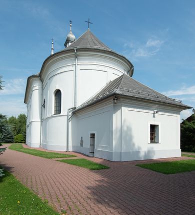 Zdjęcie nr 24: Murowany kościół otrzymał układ jednonawowej świątyni z węższym prezbiterium zwróconym ku północy i wieżą dostawioną do nawy od południa. Prezbiterium jest jednoprzęsłowe zakończone spłaszczoną apsydą. Na jego przedłużeniu usytuowano czworoboczną zakrystię. Prostokątna nawa jest trójprzęsłowa o zaokrąglonych narożnikach. Od frontu wzniesiono czworoboczną wieżę o ściętych narożnikach, flankowaną piętrowymi lokalnościami. Wysunięto ją lekko ku przodowi, gdzie przechodzi wklęsłymi płaszczyznami w boczne części fasady. Jej górne piętro nakryte cebulastym hełmem zostało opięte po bokach wolutami, obok których ustawiono kamienne klasycystyczne wazy. Elewacje zewnętrzne artykułowano podziałami ramowymi, zamykając wysokości ścian gzymsem gurtowy. Na końcu nawy na siodłowym dachu pokrytym blachą umieszczono czworoboczną wieżyczkę na sygnaturkę zwieńczoną cebulastym hełmem z krzyżem. Podobnym dachem nakryto prezbiterium. Wnętrze kościoła zostało rozczłonkowane lizenami z wklęsłymi półlizenami po bokach, ponad którymi rozciągnięto odcinki belkowania. Całość nakryto sklepieniem żaglastym na zdwojonych gurtach. Od południa w nawie urządzono chór muzyczny wsparty na dwóch kolumnach z parapetem wklęsłowypukłym. W prezbiterium usytuowano ołtarz główny, a w nawie dwa ołtarze boczne. Tu także na ścianie zachodniej zawieszono ambonę.  