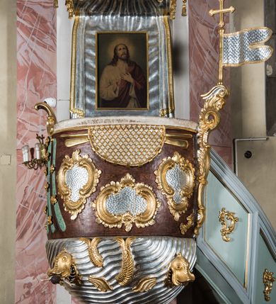 Zdjęcie nr 3: Rokokowa ambona przyścienna uzyskała kształt ożaglowanego okrętu. Składa się z kosza w formie beczkowej łodzi oraz zaplecka w kształcie żagla przechodzącego w baldachim. Kosz został podzielony na dwie strefy. W dolnej przedstawiono stylizowane, srebrzone fale morskie z igrającymi w nich morskimi, złoconymi stworami. Jeden z nich połyka postać Jonasza. Natomiast górną strefę udekorowano złoconymi, rokokowymi kartuszami, skrzyżowanymi dwoma wiosłami okrytymi fragmentem sieci rybackiej na brązowym tle. Z prawej strony do kosza przymocowano złoconą wolutę zdobioną motywem rocalliowym przechodzącym w srebrzony sztandar przymocowany do złotej laski krzyżowej. W zaplecek ambony w kształcie srebrzonego, rozpostartego żagla wstawiono nowy obraz w kształcie stojącego prostokąta, ukazujący błogosławiącego Chrystusa. Zbawiciel został przedstawiony frontalnie w dwóch trzecich postaci z głową i spojrzeniem skierowanym w górę. Prawą ręką błogosławi, lewą zaś przyciska do piersi. Jezus ma pociągłą twarz o wydatnym nosie i ustach, otoczoną brodą i długimi włosami spływającymi na ramiona. Ubrany jest w białą tunikę oraz w przerzucony przez ramię czerwony płaszcz. Jego głowę okala świetlisty nimb na ciemnym tle. Powyżej rozpięty jest baldachim utworzony z masztu podtrzymywanego przez liny i zwisających żagli zakończonych motywem lambrekinowym. Na baldachimie stoi postać uskrzydlonego anioła w luźnej, niebieskiej tunice. Ponad nim na złoconym maszcie umieszczone dwie korony, jedna nad drugą. Na jego szczycie znajduje się gloria z Duchem Świętym w postaci gołębicy na tle srebrzystych obłoków i złotych promieni. Od wschodu do ambony przylegają schody z pełną balustradą artykułowaną romboidalnym płycinami o złoconych krawędziach z polem wypełnionym złoconym motywem rocalliowym. 