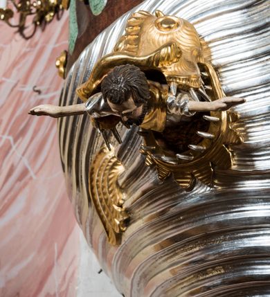 Zdjęcie nr 8: Rokokowa ambona przyścienna uzyskała kształt ożaglowanego okrętu. Składa się z kosza w formie beczkowej łodzi oraz zaplecka w kształcie żagla przechodzącego w baldachim. Kosz został podzielony na dwie strefy. W dolnej przedstawiono stylizowane, srebrzone fale morskie z igrającymi w nich morskimi, złoconymi stworami. Jeden z nich połyka postać Jonasza. Natomiast górną strefę udekorowano złoconymi, rokokowymi kartuszami, skrzyżowanymi dwoma wiosłami okrytymi fragmentem sieci rybackiej na brązowym tle. Z prawej strony do kosza przymocowano złoconą wolutę zdobioną motywem rocalliowym przechodzącym w srebrzony sztandar przymocowany do złotej laski krzyżowej. W zaplecek ambony w kształcie srebrzonego, rozpostartego żagla wstawiono nowy obraz w kształcie stojącego prostokąta, ukazujący błogosławiącego Chrystusa. Zbawiciel został przedstawiony frontalnie w dwóch trzecich postaci z głową i spojrzeniem skierowanym w górę. Prawą ręką błogosławi, lewą zaś przyciska do piersi. Jezus ma pociągłą twarz o wydatnym nosie i ustach, otoczoną brodą i długimi włosami spływającymi na ramiona. Ubrany jest w białą tunikę oraz w przerzucony przez ramię czerwony płaszcz. Jego głowę okala świetlisty nimb na ciemnym tle. Powyżej rozpięty jest baldachim utworzony z masztu podtrzymywanego przez liny i zwisających żagli zakończonych motywem lambrekinowym. Na baldachimie stoi postać uskrzydlonego anioła w luźnej, niebieskiej tunice. Ponad nim na złoconym maszcie umieszczone dwie korony, jedna nad drugą. Na jego szczycie znajduje się gloria z Duchem Świętym w postaci gołębicy na tle srebrzystych obłoków i złotych promieni. Od wschodu do ambony przylegają schody z pełną balustradą artykułowaną romboidalnym płycinami o złoconych krawędziach z polem wypełnionym złoconym motywem rocalliowym. 