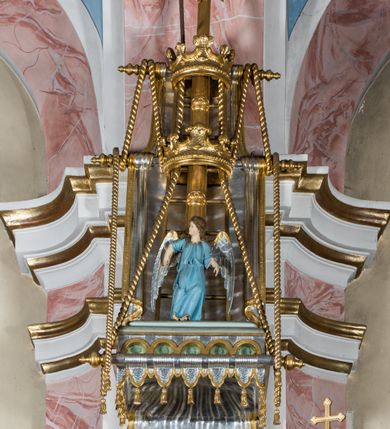 Zdjęcie nr 5: Rokokowa ambona przyścienna uzyskała kształt ożaglowanego okrętu. Składa się z kosza w formie beczkowej łodzi oraz zaplecka w kształcie żagla przechodzącego w baldachim. Kosz został podzielony na dwie strefy. W dolnej przedstawiono stylizowane, srebrzone fale morskie z igrającymi w nich morskimi, złoconymi stworami. Jeden z nich połyka postać Jonasza. Natomiast górną strefę udekorowano złoconymi, rokokowymi kartuszami, skrzyżowanymi dwoma wiosłami okrytymi fragmentem sieci rybackiej na brązowym tle. Z prawej strony do kosza przymocowano złoconą wolutę zdobioną motywem rocalliowym przechodzącym w srebrzony sztandar przymocowany do złotej laski krzyżowej. W zaplecek ambony w kształcie srebrzonego, rozpostartego żagla wstawiono nowy obraz w kształcie stojącego prostokąta, ukazujący błogosławiącego Chrystusa. Zbawiciel został przedstawiony frontalnie w dwóch trzecich postaci z głową i spojrzeniem skierowanym w górę. Prawą ręką błogosławi, lewą zaś przyciska do piersi. Jezus ma pociągłą twarz o wydatnym nosie i ustach, otoczoną brodą i długimi włosami spływającymi na ramiona. Ubrany jest w białą tunikę oraz w przerzucony przez ramię czerwony płaszcz. Jego głowę okala świetlisty nimb na ciemnym tle. Powyżej rozpięty jest baldachim utworzony z masztu podtrzymywanego przez liny i zwisających żagli zakończonych motywem lambrekinowym. Na baldachimie stoi postać uskrzydlonego anioła w luźnej, niebieskiej tunice. Ponad nim na złoconym maszcie umieszczone dwie korony, jedna nad drugą. Na jego szczycie znajduje się gloria z Duchem Świętym w postaci gołębicy na tle srebrzystych obłoków i złotych promieni. Od wschodu do ambony przylegają schody z pełną balustradą artykułowaną romboidalnym płycinami o złoconych krawędziach z polem wypełnionym złoconym motywem rocalliowym. 