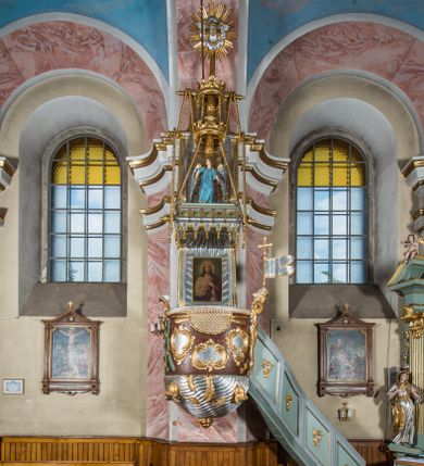 Zdjęcie nr 1: Rokokowa ambona przyścienna uzyskała kształt ożaglowanego okrętu. Składa się z kosza w formie beczkowej łodzi oraz zaplecka w kształcie żagla przechodzącego w baldachim. Kosz został podzielony na dwie strefy. W dolnej przedstawiono stylizowane, srebrzone fale morskie z igrającymi w nich morskimi, złoconymi stworami. Jeden z nich połyka postać Jonasza. Natomiast górną strefę udekorowano złoconymi, rokokowymi kartuszami, skrzyżowanymi dwoma wiosłami okrytymi fragmentem sieci rybackiej na brązowym tle. Z prawej strony do kosza przymocowano złoconą wolutę zdobioną motywem rocalliowym przechodzącym w srebrzony sztandar przymocowany do złotej laski krzyżowej. W zaplecek ambony w kształcie srebrzonego, rozpostartego żagla wstawiono nowy obraz w kształcie stojącego prostokąta, ukazujący błogosławiącego Chrystusa. Zbawiciel został przedstawiony frontalnie w dwóch trzecich postaci z głową i spojrzeniem skierowanym w górę. Prawą ręką błogosławi, lewą zaś przyciska do piersi. Jezus ma pociągłą twarz o wydatnym nosie i ustach, otoczoną brodą i długimi włosami spływającymi na ramiona. Ubrany jest w białą tunikę oraz w przerzucony przez ramię czerwony płaszcz. Jego głowę okala świetlisty nimb na ciemnym tle. Powyżej rozpięty jest baldachim utworzony z masztu podtrzymywanego przez liny i zwisających żagli zakończonych motywem lambrekinowym. Na baldachimie stoi postać uskrzydlonego anioła w luźnej, niebieskiej tunice. Ponad nim na złoconym maszcie umieszczone dwie korony, jedna nad drugą. Na jego szczycie znajduje się gloria z Duchem Świętym w postaci gołębicy na tle srebrzystych obłoków i złotych promieni. Od wschodu do ambony przylegają schody z pełną balustradą artykułowaną romboidalnym płycinami o złoconych krawędziach z polem wypełnionym złoconym motywem rocalliowym. 