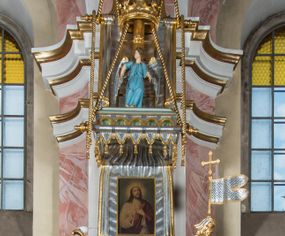 Zdjęcie nr 1: Rokokowa ambona przyścienna uzyskała kształt ożaglowanego okrętu. Składa się z kosza w formie beczkowej łodzi oraz zaplecka w kształcie żagla przechodzącego w baldachim. Kosz został podzielony na dwie strefy. W dolnej przedstawiono stylizowane, srebrzone fale morskie z igrającymi w nich morskimi, złoconymi stworami. Jeden z nich połyka postać Jonasza. Natomiast górną strefę udekorowano złoconymi, rokokowymi kartuszami, skrzyżowanymi dwoma wiosłami okrytymi fragmentem sieci rybackiej na brązowym tle. Z prawej strony do kosza przymocowano złoconą wolutę zdobioną motywem rocalliowym przechodzącym w srebrzony sztandar przymocowany do złotej laski krzyżowej. W zaplecek ambony w kształcie srebrzonego, rozpostartego żagla wstawiono nowy obraz w kształcie stojącego prostokąta, ukazujący błogosławiącego Chrystusa. Zbawiciel został przedstawiony frontalnie w dwóch trzecich postaci z głową i spojrzeniem skierowanym w górę. Prawą ręką błogosławi, lewą zaś przyciska do piersi. Jezus ma pociągłą twarz o wydatnym nosie i ustach, otoczoną brodą i długimi włosami spływającymi na ramiona. Ubrany jest w białą tunikę oraz w przerzucony przez ramię czerwony płaszcz. Jego głowę okala świetlisty nimb na ciemnym tle. Powyżej rozpięty jest baldachim utworzony z masztu podtrzymywanego przez liny i zwisających żagli zakończonych motywem lambrekinowym. Na baldachimie stoi postać uskrzydlonego anioła w luźnej, niebieskiej tunice. Ponad nim na złoconym maszcie umieszczone dwie korony, jedna nad drugą. Na jego szczycie znajduje się gloria z Duchem Świętym w postaci gołębicy na tle srebrzystych obłoków i złotych promieni. Od wschodu do ambony przylegają schody z pełną balustradą artykułowaną romboidalnym płycinami o złoconych krawędziach z polem wypełnionym złoconym motywem rocalliowym. 