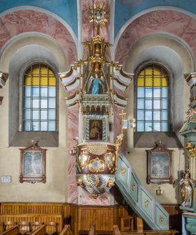 Zdjęcie nr 1: Rokokowa ambona przyścienna uzyskała kształt ożaglowanego okrętu. Składa się z kosza w formie beczkowej łodzi oraz zaplecka w kształcie żagla przechodzącego w baldachim. Kosz został podzielony na dwie strefy. W dolnej przedstawiono stylizowane, srebrzone fale morskie z igrającymi w nich morskimi, złoconymi stworami. Jeden z nich połyka postać Jonasza. Natomiast górną strefę udekorowano złoconymi, rokokowymi kartuszami, skrzyżowanymi dwoma wiosłami okrytymi fragmentem sieci rybackiej na brązowym tle. Z prawej strony do kosza przymocowano złoconą wolutę zdobioną motywem rocalliowym przechodzącym w srebrzony sztandar przymocowany do złotej laski krzyżowej. W zaplecek ambony w kształcie srebrzonego, rozpostartego żagla wstawiono nowy obraz w kształcie stojącego prostokąta, ukazujący błogosławiącego Chrystusa. Zbawiciel został przedstawiony frontalnie w dwóch trzecich postaci z głową i spojrzeniem skierowanym w górę. Prawą ręką błogosławi, lewą zaś przyciska do piersi. Jezus ma pociągłą twarz o wydatnym nosie i ustach, otoczoną brodą i długimi włosami spływającymi na ramiona. Ubrany jest w białą tunikę oraz w przerzucony przez ramię czerwony płaszcz. Jego głowę okala świetlisty nimb na ciemnym tle. Powyżej rozpięty jest baldachim utworzony z masztu podtrzymywanego przez liny i zwisających żagli zakończonych motywem lambrekinowym. Na baldachimie stoi postać uskrzydlonego anioła w luźnej, niebieskiej tunice. Ponad nim na złoconym maszcie umieszczone dwie korony, jedna nad drugą. Na jego szczycie znajduje się gloria z Duchem Świętym w postaci gołębicy na tle srebrzystych obłoków i złotych promieni. Od wschodu do ambony przylegają schody z pełną balustradą artykułowaną romboidalnym płycinami o złoconych krawędziach z polem wypełnionym złoconym motywem rocalliowym. 