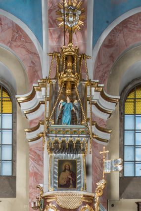 Zdjęcie nr 1: Rokokowa ambona przyścienna uzyskała kształt ożaglowanego okrętu. Składa się z kosza w formie beczkowej łodzi oraz zaplecka w kształcie żagla przechodzącego w baldachim. Kosz został podzielony na dwie strefy. W dolnej przedstawiono stylizowane, srebrzone fale morskie z igrającymi w nich morskimi, złoconymi stworami. Jeden z nich połyka postać Jonasza. Natomiast górną strefę udekorowano złoconymi, rokokowymi kartuszami, skrzyżowanymi dwoma wiosłami okrytymi fragmentem sieci rybackiej na brązowym tle. Z prawej strony do kosza przymocowano złoconą wolutę zdobioną motywem rocalliowym przechodzącym w srebrzony sztandar przymocowany do złotej laski krzyżowej. W zaplecek ambony w kształcie srebrzonego, rozpostartego żagla wstawiono nowy obraz w kształcie stojącego prostokąta, ukazujący błogosławiącego Chrystusa. Zbawiciel został przedstawiony frontalnie w dwóch trzecich postaci z głową i spojrzeniem skierowanym w górę. Prawą ręką błogosławi, lewą zaś przyciska do piersi. Jezus ma pociągłą twarz o wydatnym nosie i ustach, otoczoną brodą i długimi włosami spływającymi na ramiona. Ubrany jest w białą tunikę oraz w przerzucony przez ramię czerwony płaszcz. Jego głowę okala świetlisty nimb na ciemnym tle. Powyżej rozpięty jest baldachim utworzony z masztu podtrzymywanego przez liny i zwisających żagli zakończonych motywem lambrekinowym. Na baldachimie stoi postać uskrzydlonego anioła w luźnej, niebieskiej tunice. Ponad nim na złoconym maszcie umieszczone dwie korony, jedna nad drugą. Na jego szczycie znajduje się gloria z Duchem Świętym w postaci gołębicy na tle srebrzystych obłoków i złotych promieni. Od wschodu do ambony przylegają schody z pełną balustradą artykułowaną romboidalnym płycinami o złoconych krawędziach z polem wypełnionym złoconym motywem rocalliowym. 