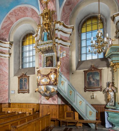 Zdjęcie nr 2: Rokokowa ambona przyścienna uzyskała kształt ożaglowanego okrętu. Składa się z kosza w formie beczkowej łodzi oraz zaplecka w kształcie żagla przechodzącego w baldachim. Kosz został podzielony na dwie strefy. W dolnej przedstawiono stylizowane, srebrzone fale morskie z igrającymi w nich morskimi, złoconymi stworami. Jeden z nich połyka postać Jonasza. Natomiast górną strefę udekorowano złoconymi, rokokowymi kartuszami, skrzyżowanymi dwoma wiosłami okrytymi fragmentem sieci rybackiej na brązowym tle. Z prawej strony do kosza przymocowano złoconą wolutę zdobioną motywem rocalliowym przechodzącym w srebrzony sztandar przymocowany do złotej laski krzyżowej. W zaplecek ambony w kształcie srebrzonego, rozpostartego żagla wstawiono nowy obraz w kształcie stojącego prostokąta, ukazujący błogosławiącego Chrystusa. Zbawiciel został przedstawiony frontalnie w dwóch trzecich postaci z głową i spojrzeniem skierowanym w górę. Prawą ręką błogosławi, lewą zaś przyciska do piersi. Jezus ma pociągłą twarz o wydatnym nosie i ustach, otoczoną brodą i długimi włosami spływającymi na ramiona. Ubrany jest w białą tunikę oraz w przerzucony przez ramię czerwony płaszcz. Jego głowę okala świetlisty nimb na ciemnym tle. Powyżej rozpięty jest baldachim utworzony z masztu podtrzymywanego przez liny i zwisających żagli zakończonych motywem lambrekinowym. Na baldachimie stoi postać uskrzydlonego anioła w luźnej, niebieskiej tunice. Ponad nim na złoconym maszcie umieszczone dwie korony, jedna nad drugą. Na jego szczycie znajduje się gloria z Duchem Świętym w postaci gołębicy na tle srebrzystych obłoków i złotych promieni. Od wschodu do ambony przylegają schody z pełną balustradą artykułowaną romboidalnym płycinami o złoconych krawędziach z polem wypełnionym złoconym motywem rocalliowym. 