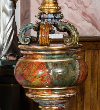 Zdjęcie nr 1: Chrzcielnica uzyskała kształt kielicha. Czaszę w formie ściętej kuli, zaopatrzoną u spodu w profilowanie, wsparto na bogato toczonym trzonie w kształcie tralki, przechodzącym w profilowaną stopę ustawioną na wielobocznym cokoliku. Płaska nakrywa chrzcielnicy  ozdobiona została połączonymi ze sobą sześcioma spływami wolutowymi, zdobionymi motywem kaboszonów, spiętymi w górze masywną, zamkniętą koroną. Całość pokrywa polichromia marmoryzowana w kolorach szaro-zielonkawych. Zwieńczenie i niektóre profile są złocone. 