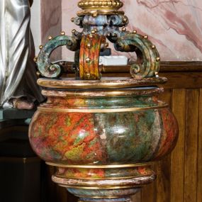 Zdjęcie nr 1: Chrzcielnica uzyskała kształt kielicha. Czaszę w formie ściętej kuli, zaopatrzoną u spodu w profilowanie, wsparto na bogato toczonym trzonie w kształcie tralki, przechodzącym w profilowaną stopę ustawioną na wielobocznym cokoliku. Płaska nakrywa chrzcielnicy  ozdobiona została połączonymi ze sobą sześcioma spływami wolutowymi, zdobionymi motywem kaboszonów, spiętymi w górze masywną, zamkniętą koroną. Całość pokrywa polichromia marmoryzowana w kolorach szaro-zielonkawych. Zwieńczenie i niektóre profile są złocone. 