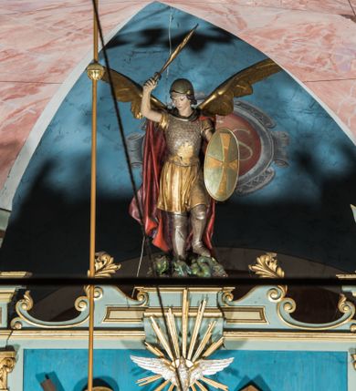 Zdjęcie nr 20: Ołtarz główny uzyskał formę przyściennej, dwukondygnacyjnej, neoklasycystycznej nastawy wzniesionej na rzucie elipsy z bocznymi bramkami. Stipes ołtarzowy uzyskał kształt leżącego prostopadłościanu podtrzymującego drewnianą mensę z portatylem. Jego lico dekorowane jest trzema równoległymi płycinami utworzonymi ze złoconych, profilowanych listew. Na mensie ustawiono prostopadłościenne tabernakulum w formie skrzynki flankowane parą pilastrów podtrzymujących nastawę tronu eucharystycznego. Ujmują go po bokach pary złoconych kolumn z korynckimi kapitelami podtrzymujące belkowanie wygięte na osi półkoliście. Na zwieńczeniu tabernakulum ustawiono leżącego na zamkniętej księdze z siedmioma pieczęciami srebrnego baranka apokaliptycznego na tle srebrzonych promieni. Pomiędzy kolumnami w polu zainstalowano obrotowy bęben z dwiema półkolistymi niszami. Retabulum zostało wsparte na wysokim cokole. Dolną kondygnację artykułują zwielokrotnione pilastry o kanelowanych trzonach i złoconych, kompozytowych głowicach oraz dwie wysunięte do przodu kanelowane kolumny o kapitelach kompozytowych. Podpory podtrzymują szeroki architraw wklęsło-wypukły dekorowany motywami roślinnymi oraz perełkami i ząbkami, wyłamany na przedłużeniu kolumn. W miejscu wyłamań usytuowano siedzących dwóch uskrzydlonych aniołów. Po bokach kolumn na konsolach ustawiono figury św. Piotra i św. Pawła. Boki ołtarza zamykają ażurowe wici roślinne pełniące rolę zredukowanych uszaków. W polu środkowym retabulum umieszczono prostokątne obramienie utworzone z dekoracyjnej ramy zdobionej wicią roślinną oraz podwieszonego u jej góry paludamentum z koroną podtrzymywaną przez dwa aniołki. W środku zainstalowano obraz Matki Boskiej Śnieżnej, a na zasuwie przedstawienie Chrztu Chrystusa w Jordanie. Zwieńczenie nastawy uzyskało kształt leżącego prostokąta flankowanego po bokach parą kanelowanych pilastrów o korynckich głowicach, na tle których ustawiono dwie kolumny podtrzymujących przerwane belkowanie z przerwanym przyczółkiem. Na osi zwieńczenia na postumencie ustawiono rzeźbę św. Michała Archanioła, a na przyczółkach rzeźby dwóch siedzących aniołów. W polu między pilastrami i kolumnami znalazło się płaskorzeźbione przedstawienie Trójcy Przenajświętszej. Dwie flankujące retabulum bramki ozdobiono dwoma aniołkami siedzącymi na ich zwieńczeniu na postumencie. Struktura całości pomalowane jest na zielono, ornamenty i profile wyzłocono, a rzeźby otrzymały naturalistyczną polichromię.