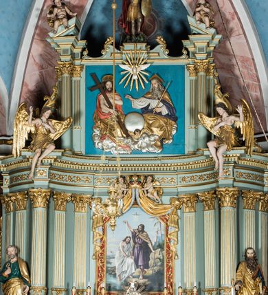 Zdjęcie nr 4: Ołtarz główny uzyskał formę przyściennej, dwukondygnacyjnej, neoklasycystycznej nastawy wzniesionej na rzucie elipsy z bocznymi bramkami. Stipes ołtarzowy uzyskał kształt leżącego prostopadłościanu podtrzymującego drewnianą mensę z portatylem. Jego lico dekorowane jest trzema równoległymi płycinami utworzonymi ze złoconych, profilowanych listew. Na mensie ustawiono prostopadłościenne tabernakulum w formie skrzynki flankowane parą pilastrów podtrzymujących nastawę tronu eucharystycznego. Ujmują go po bokach pary złoconych kolumn z korynckimi kapitelami podtrzymujące belkowanie wygięte na osi półkoliście. Na zwieńczeniu tabernakulum ustawiono leżącego na zamkniętej księdze z siedmioma pieczęciami srebrnego baranka apokaliptycznego na tle srebrzonych promieni. Pomiędzy kolumnami w polu zainstalowano obrotowy bęben z dwiema półkolistymi niszami. Retabulum zostało wsparte na wysokim cokole. Dolną kondygnację artykułują zwielokrotnione pilastry o kanelowanych trzonach i złoconych, kompozytowych głowicach oraz dwie wysunięte do przodu kanelowane kolumny o kapitelach kompozytowych. Podpory podtrzymują szeroki architraw wklęsło-wypukły dekorowany motywami roślinnymi oraz perełkami i ząbkami, wyłamany na przedłużeniu kolumn. W miejscu wyłamań usytuowano siedzących dwóch uskrzydlonych aniołów. Po bokach kolumn na konsolach ustawiono figury św. Piotra i św. Pawła. Boki ołtarza zamykają ażurowe wici roślinne pełniące rolę zredukowanych uszaków. W polu środkowym retabulum umieszczono prostokątne obramienie utworzone z dekoracyjnej ramy zdobionej wicią roślinną oraz podwieszonego u jej góry paludamentum z koroną podtrzymywaną przez dwa aniołki. W środku zainstalowano obraz Matki Boskiej Śnieżnej, a na zasuwie przedstawienie Chrztu Chrystusa w Jordanie. Zwieńczenie nastawy uzyskało kształt leżącego prostokąta flankowanego po bokach parą kanelowanych pilastrów o korynckich głowicach, na tle których ustawiono dwie kolumny podtrzymujących przerwane belkowanie z przerwanym przyczółkiem. Na osi zwieńczenia na postumencie ustawiono rzeźbę św. Michała Archanioła, a na przyczółkach rzeźby dwóch siedzących aniołów. W polu między pilastrami i kolumnami znalazło się płaskorzeźbione przedstawienie Trójcy Przenajświętszej. Dwie flankujące retabulum bramki ozdobiono dwoma aniołkami siedzącymi na ich zwieńczeniu na postumencie. Struktura całości pomalowane jest na zielono, ornamenty i profile wyzłocono, a rzeźby otrzymały naturalistyczną polichromię.