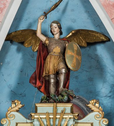 Zdjęcie nr 25: Ołtarz główny uzyskał formę przyściennej, dwukondygnacyjnej, neoklasycystycznej nastawy wzniesionej na rzucie elipsy z bocznymi bramkami. Stipes ołtarzowy uzyskał kształt leżącego prostopadłościanu podtrzymującego drewnianą mensę z portatylem. Jego lico dekorowane jest trzema równoległymi płycinami utworzonymi ze złoconych, profilowanych listew. Na mensie ustawiono prostopadłościenne tabernakulum w formie skrzynki flankowane parą pilastrów podtrzymujących nastawę tronu eucharystycznego. Ujmują go po bokach pary złoconych kolumn z korynckimi kapitelami podtrzymujące belkowanie wygięte na osi półkoliście. Na zwieńczeniu tabernakulum ustawiono leżącego na zamkniętej księdze z siedmioma pieczęciami srebrnego baranka apokaliptycznego na tle srebrzonych promieni. Pomiędzy kolumnami w polu zainstalowano obrotowy bęben z dwiema półkolistymi niszami. Retabulum zostało wsparte na wysokim cokole. Dolną kondygnację artykułują zwielokrotnione pilastry o kanelowanych trzonach i złoconych, kompozytowych głowicach oraz dwie wysunięte do przodu kanelowane kolumny o kapitelach kompozytowych. Podpory podtrzymują szeroki architraw wklęsło-wypukły dekorowany motywami roślinnymi oraz perełkami i ząbkami, wyłamany na przedłużeniu kolumn. W miejscu wyłamań usytuowano siedzących dwóch uskrzydlonych aniołów. Po bokach kolumn na konsolach ustawiono figury św. Piotra i św. Pawła. Boki ołtarza zamykają ażurowe wici roślinne pełniące rolę zredukowanych uszaków. W polu środkowym retabulum umieszczono prostokątne obramienie utworzone z dekoracyjnej ramy zdobionej wicią roślinną oraz podwieszonego u jej góry paludamentum z koroną podtrzymywaną przez dwa aniołki. W środku zainstalowano obraz Matki Boskiej Śnieżnej, a na zasuwie przedstawienie Chrztu Chrystusa w Jordanie. Zwieńczenie nastawy uzyskało kształt leżącego prostokąta flankowanego po bokach parą kanelowanych pilastrów o korynckich głowicach, na tle których ustawiono dwie kolumny podtrzymujących przerwane belkowanie z przerwanym przyczółkiem. Na osi zwieńczenia na postumencie ustawiono rzeźbę św. Michała Archanioła, a na przyczółkach rzeźby dwóch siedzących aniołów. W polu między pilastrami i kolumnami znalazło się płaskorzeźbione przedstawienie Trójcy Przenajświętszej. Dwie flankujące retabulum bramki ozdobiono dwoma aniołkami siedzącymi na ich zwieńczeniu na postumencie. Struktura całości pomalowane jest na zielono, ornamenty i profile wyzłocono, a rzeźby otrzymały naturalistyczną polichromię.