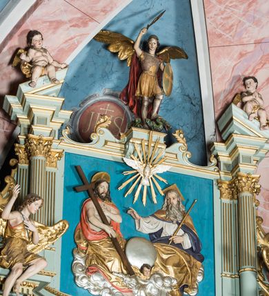 Zdjęcie nr 13: Ołtarz główny uzyskał formę przyściennej, dwukondygnacyjnej, neoklasycystycznej nastawy wzniesionej na rzucie elipsy z bocznymi bramkami. Stipes ołtarzowy uzyskał kształt leżącego prostopadłościanu podtrzymującego drewnianą mensę z portatylem. Jego lico dekorowane jest trzema równoległymi płycinami utworzonymi ze złoconych, profilowanych listew. Na mensie ustawiono prostopadłościenne tabernakulum w formie skrzynki flankowane parą pilastrów podtrzymujących nastawę tronu eucharystycznego. Ujmują go po bokach pary złoconych kolumn z korynckimi kapitelami podtrzymujące belkowanie wygięte na osi półkoliście. Na zwieńczeniu tabernakulum ustawiono leżącego na zamkniętej księdze z siedmioma pieczęciami srebrnego baranka apokaliptycznego na tle srebrzonych promieni. Pomiędzy kolumnami w polu zainstalowano obrotowy bęben z dwiema półkolistymi niszami. Retabulum zostało wsparte na wysokim cokole. Dolną kondygnację artykułują zwielokrotnione pilastry o kanelowanych trzonach i złoconych, kompozytowych głowicach oraz dwie wysunięte do przodu kanelowane kolumny o kapitelach kompozytowych. Podpory podtrzymują szeroki architraw wklęsło-wypukły dekorowany motywami roślinnymi oraz perełkami i ząbkami, wyłamany na przedłużeniu kolumn. W miejscu wyłamań usytuowano siedzących dwóch uskrzydlonych aniołów. Po bokach kolumn na konsolach ustawiono figury św. Piotra i św. Pawła. Boki ołtarza zamykają ażurowe wici roślinne pełniące rolę zredukowanych uszaków. W polu środkowym retabulum umieszczono prostokątne obramienie utworzone z dekoracyjnej ramy zdobionej wicią roślinną oraz podwieszonego u jej góry paludamentum z koroną podtrzymywaną przez dwa aniołki. W środku zainstalowano obraz Matki Boskiej Śnieżnej, a na zasuwie przedstawienie Chrztu Chrystusa w Jordanie. Zwieńczenie nastawy uzyskało kształt leżącego prostokąta flankowanego po bokach parą kanelowanych pilastrów o korynckich głowicach, na tle których ustawiono dwie kolumny podtrzymujących przerwane belkowanie z przerwanym przyczółkiem. Na osi zwieńczenia na postumencie ustawiono rzeźbę św. Michała Archanioła, a na przyczółkach rzeźby dwóch siedzących aniołów. W polu między pilastrami i kolumnami znalazło się płaskorzeźbione przedstawienie Trójcy Przenajświętszej. Dwie flankujące retabulum bramki ozdobiono dwoma aniołkami siedzącymi na ich zwieńczeniu na postumencie. Struktura całości pomalowane jest na zielono, ornamenty i profile wyzłocono, a rzeźby otrzymały naturalistyczną polichromię.