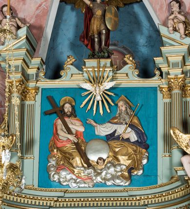 Zdjęcie nr 14: Ołtarz główny uzyskał formę przyściennej, dwukondygnacyjnej, neoklasycystycznej nastawy wzniesionej na rzucie elipsy z bocznymi bramkami. Stipes ołtarzowy uzyskał kształt leżącego prostopadłościanu podtrzymującego drewnianą mensę z portatylem. Jego lico dekorowane jest trzema równoległymi płycinami utworzonymi ze złoconych, profilowanych listew. Na mensie ustawiono prostopadłościenne tabernakulum w formie skrzynki flankowane parą pilastrów podtrzymujących nastawę tronu eucharystycznego. Ujmują go po bokach pary złoconych kolumn z korynckimi kapitelami podtrzymujące belkowanie wygięte na osi półkoliście. Na zwieńczeniu tabernakulum ustawiono leżącego na zamkniętej księdze z siedmioma pieczęciami srebrnego baranka apokaliptycznego na tle srebrzonych promieni. Pomiędzy kolumnami w polu zainstalowano obrotowy bęben z dwiema półkolistymi niszami. Retabulum zostało wsparte na wysokim cokole. Dolną kondygnację artykułują zwielokrotnione pilastry o kanelowanych trzonach i złoconych, kompozytowych głowicach oraz dwie wysunięte do przodu kanelowane kolumny o kapitelach kompozytowych. Podpory podtrzymują szeroki architraw wklęsło-wypukły dekorowany motywami roślinnymi oraz perełkami i ząbkami, wyłamany na przedłużeniu kolumn. W miejscu wyłamań usytuowano siedzących dwóch uskrzydlonych aniołów. Po bokach kolumn na konsolach ustawiono figury św. Piotra i św. Pawła. Boki ołtarza zamykają ażurowe wici roślinne pełniące rolę zredukowanych uszaków. W polu środkowym retabulum umieszczono prostokątne obramienie utworzone z dekoracyjnej ramy zdobionej wicią roślinną oraz podwieszonego u jej góry paludamentum z koroną podtrzymywaną przez dwa aniołki. W środku zainstalowano obraz Matki Boskiej Śnieżnej, a na zasuwie przedstawienie Chrztu Chrystusa w Jordanie. Zwieńczenie nastawy uzyskało kształt leżącego prostokąta flankowanego po bokach parą kanelowanych pilastrów o korynckich głowicach, na tle których ustawiono dwie kolumny podtrzymujących przerwane belkowanie z przerwanym przyczółkiem. Na osi zwieńczenia na postumencie ustawiono rzeźbę św. Michała Archanioła, a na przyczółkach rzeźby dwóch siedzących aniołów. W polu między pilastrami i kolumnami znalazło się płaskorzeźbione przedstawienie Trójcy Przenajświętszej. Dwie flankujące retabulum bramki ozdobiono dwoma aniołkami siedzącymi na ich zwieńczeniu na postumencie. Struktura całości pomalowane jest na zielono, ornamenty i profile wyzłocono, a rzeźby otrzymały naturalistyczną polichromię.