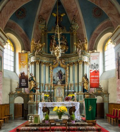Zdjęcie nr 1: Ołtarz główny uzyskał formę przyściennej, dwukondygnacyjnej, neoklasycystycznej nastawy wzniesionej na rzucie elipsy z bocznymi bramkami. Stipes ołtarzowy uzyskał kształt leżącego prostopadłościanu podtrzymującego drewnianą mensę z portatylem. Jego lico dekorowane jest trzema równoległymi płycinami utworzonymi ze złoconych, profilowanych listew. Na mensie ustawiono prostopadłościenne tabernakulum w formie skrzynki flankowane parą pilastrów podtrzymujących nastawę tronu eucharystycznego. Ujmują go po bokach pary złoconych kolumn z korynckimi kapitelami podtrzymujące belkowanie wygięte na osi półkoliście. Na zwieńczeniu tabernakulum ustawiono leżącego na zamkniętej księdze z siedmioma pieczęciami srebrnego baranka apokaliptycznego na tle srebrzonych promieni. Pomiędzy kolumnami w polu zainstalowano obrotowy bęben z dwiema półkolistymi niszami. Retabulum zostało wsparte na wysokim cokole. Dolną kondygnację artykułują zwielokrotnione pilastry o kanelowanych trzonach i złoconych, kompozytowych głowicach oraz dwie wysunięte do przodu kanelowane kolumny o kapitelach kompozytowych. Podpory podtrzymują szeroki architraw wklęsło-wypukły dekorowany motywami roślinnymi oraz perełkami i ząbkami, wyłamany na przedłużeniu kolumn. W miejscu wyłamań usytuowano siedzących dwóch uskrzydlonych aniołów. Po bokach kolumn na konsolach ustawiono figury św. Piotra i św. Pawła. Boki ołtarza zamykają ażurowe wici roślinne pełniące rolę zredukowanych uszaków. W polu środkowym retabulum umieszczono prostokątne obramienie utworzone z dekoracyjnej ramy zdobionej wicią roślinną oraz podwieszonego u jej góry paludamentum z koroną podtrzymywaną przez dwa aniołki. W środku zainstalowano obraz Matki Boskiej Śnieżnej, a na zasuwie przedstawienie Chrztu Chrystusa w Jordanie. Zwieńczenie nastawy uzyskało kształt leżącego prostokąta flankowanego po bokach parą kanelowanych pilastrów o korynckich głowicach, na tle których ustawiono dwie kolumny podtrzymujących przerwane belkowanie z przerwanym przyczółkiem. Na osi zwieńczenia na postumencie ustawiono rzeźbę św. Michała Archanioła, a na przyczółkach rzeźby dwóch siedzących aniołów. W polu między pilastrami i kolumnami znalazło się płaskorzeźbione przedstawienie Trójcy Przenajświętszej. Dwie flankujące retabulum bramki ozdobiono dwoma aniołkami siedzącymi na ich zwieńczeniu na postumencie. Struktura całości pomalowane jest na zielono, ornamenty i profile wyzłocono, a rzeźby otrzymały naturalistyczną polichromię.