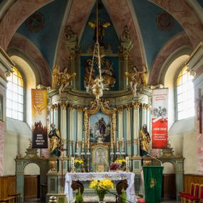 Zdjęcie nr 1: Ołtarz główny uzyskał formę przyściennej, dwukondygnacyjnej, neoklasycystycznej nastawy wzniesionej na rzucie elipsy z bocznymi bramkami. Stipes ołtarzowy uzyskał kształt leżącego prostopadłościanu podtrzymującego drewnianą mensę z portatylem. Jego lico dekorowane jest trzema równoległymi płycinami utworzonymi ze złoconych, profilowanych listew. Na mensie ustawiono prostopadłościenne tabernakulum w formie skrzynki flankowane parą pilastrów podtrzymujących nastawę tronu eucharystycznego. Ujmują go po bokach pary złoconych kolumn z korynckimi kapitelami podtrzymujące belkowanie wygięte na osi półkoliście. Na zwieńczeniu tabernakulum ustawiono leżącego na zamkniętej księdze z siedmioma pieczęciami srebrnego baranka apokaliptycznego na tle srebrzonych promieni. Pomiędzy kolumnami w polu zainstalowano obrotowy bęben z dwiema półkolistymi niszami. Retabulum zostało wsparte na wysokim cokole. Dolną kondygnację artykułują zwielokrotnione pilastry o kanelowanych trzonach i złoconych, kompozytowych głowicach oraz dwie wysunięte do przodu kanelowane kolumny o kapitelach kompozytowych. Podpory podtrzymują szeroki architraw wklęsło-wypukły dekorowany motywami roślinnymi oraz perełkami i ząbkami, wyłamany na przedłużeniu kolumn. W miejscu wyłamań usytuowano siedzących dwóch uskrzydlonych aniołów. Po bokach kolumn na konsolach ustawiono figury św. Piotra i św. Pawła. Boki ołtarza zamykają ażurowe wici roślinne pełniące rolę zredukowanych uszaków. W polu środkowym retabulum umieszczono prostokątne obramienie utworzone z dekoracyjnej ramy zdobionej wicią roślinną oraz podwieszonego u jej góry paludamentum z koroną podtrzymywaną przez dwa aniołki. W środku zainstalowano obraz Matki Boskiej Śnieżnej, a na zasuwie przedstawienie Chrztu Chrystusa w Jordanie. Zwieńczenie nastawy uzyskało kształt leżącego prostokąta flankowanego po bokach parą kanelowanych pilastrów o korynckich głowicach, na tle których ustawiono dwie kolumny podtrzymujących przerwane belkowanie z przerwanym przyczółkiem. Na osi zwieńczenia na postumencie ustawiono rzeźbę św. Michała Archanioła, a na przyczółkach rzeźby dwóch siedzących aniołów. W polu między pilastrami i kolumnami znalazło się płaskorzeźbione przedstawienie Trójcy Przenajświętszej. Dwie flankujące retabulum bramki ozdobiono dwoma aniołkami siedzącymi na ich zwieńczeniu na postumencie. Struktura całości pomalowane jest na zielono, ornamenty i profile wyzłocono, a rzeźby otrzymały naturalistyczną polichromię.