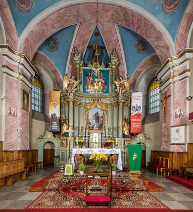 Zdjęcie nr 2: Ołtarz główny uzyskał formę przyściennej, dwukondygnacyjnej, neoklasycystycznej nastawy wzniesionej na rzucie elipsy z bocznymi bramkami. Stipes ołtarzowy uzyskał kształt leżącego prostopadłościanu podtrzymującego drewnianą mensę z portatylem. Jego lico dekorowane jest trzema równoległymi płycinami utworzonymi ze złoconych, profilowanych listew. Na mensie ustawiono prostopadłościenne tabernakulum w formie skrzynki flankowane parą pilastrów podtrzymujących nastawę tronu eucharystycznego. Ujmują go po bokach pary złoconych kolumn z korynckimi kapitelami podtrzymujące belkowanie wygięte na osi półkoliście. Na zwieńczeniu tabernakulum ustawiono leżącego na zamkniętej księdze z siedmioma pieczęciami srebrnego baranka apokaliptycznego na tle srebrzonych promieni. Pomiędzy kolumnami w polu zainstalowano obrotowy bęben z dwiema półkolistymi niszami. Retabulum zostało wsparte na wysokim cokole. Dolną kondygnację artykułują zwielokrotnione pilastry o kanelowanych trzonach i złoconych, kompozytowych głowicach oraz dwie wysunięte do przodu kanelowane kolumny o kapitelach kompozytowych. Podpory podtrzymują szeroki architraw wklęsło-wypukły dekorowany motywami roślinnymi oraz perełkami i ząbkami, wyłamany na przedłużeniu kolumn. W miejscu wyłamań usytuowano siedzących dwóch uskrzydlonych aniołów. Po bokach kolumn na konsolach ustawiono figury św. Piotra i św. Pawła. Boki ołtarza zamykają ażurowe wici roślinne pełniące rolę zredukowanych uszaków. W polu środkowym retabulum umieszczono prostokątne obramienie utworzone z dekoracyjnej ramy zdobionej wicią roślinną oraz podwieszonego u jej góry paludamentum z koroną podtrzymywaną przez dwa aniołki. W środku zainstalowano obraz Matki Boskiej Śnieżnej, a na zasuwie przedstawienie Chrztu Chrystusa w Jordanie. Zwieńczenie nastawy uzyskało kształt leżącego prostokąta flankowanego po bokach parą kanelowanych pilastrów o korynckich głowicach, na tle których ustawiono dwie kolumny podtrzymujących przerwane belkowanie z przerwanym przyczółkiem. Na osi zwieńczenia na postumencie ustawiono rzeźbę św. Michała Archanioła, a na przyczółkach rzeźby dwóch siedzących aniołów. W polu między pilastrami i kolumnami znalazło się płaskorzeźbione przedstawienie Trójcy Przenajświętszej. Dwie flankujące retabulum bramki ozdobiono dwoma aniołkami siedzącymi na ich zwieńczeniu na postumencie. Struktura całości pomalowane jest na zielono, ornamenty i profile wyzłocono, a rzeźby otrzymały naturalistyczną polichromię.