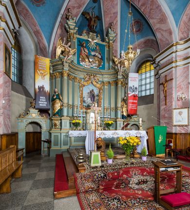 Zdjęcie nr 3: Ołtarz główny uzyskał formę przyściennej, dwukondygnacyjnej, neoklasycystycznej nastawy wzniesionej na rzucie elipsy z bocznymi bramkami. Stipes ołtarzowy uzyskał kształt leżącego prostopadłościanu podtrzymującego drewnianą mensę z portatylem. Jego lico dekorowane jest trzema równoległymi płycinami utworzonymi ze złoconych, profilowanych listew. Na mensie ustawiono prostopadłościenne tabernakulum w formie skrzynki flankowane parą pilastrów podtrzymujących nastawę tronu eucharystycznego. Ujmują go po bokach pary złoconych kolumn z korynckimi kapitelami podtrzymujące belkowanie wygięte na osi półkoliście. Na zwieńczeniu tabernakulum ustawiono leżącego na zamkniętej księdze z siedmioma pieczęciami srebrnego baranka apokaliptycznego na tle srebrzonych promieni. Pomiędzy kolumnami w polu zainstalowano obrotowy bęben z dwiema półkolistymi niszami. Retabulum zostało wsparte na wysokim cokole. Dolną kondygnację artykułują zwielokrotnione pilastry o kanelowanych trzonach i złoconych, kompozytowych głowicach oraz dwie wysunięte do przodu kanelowane kolumny o kapitelach kompozytowych. Podpory podtrzymują szeroki architraw wklęsło-wypukły dekorowany motywami roślinnymi oraz perełkami i ząbkami, wyłamany na przedłużeniu kolumn. W miejscu wyłamań usytuowano siedzących dwóch uskrzydlonych aniołów. Po bokach kolumn na konsolach ustawiono figury św. Piotra i św. Pawła. Boki ołtarza zamykają ażurowe wici roślinne pełniące rolę zredukowanych uszaków. W polu środkowym retabulum umieszczono prostokątne obramienie utworzone z dekoracyjnej ramy zdobionej wicią roślinną oraz podwieszonego u jej góry paludamentum z koroną podtrzymywaną przez dwa aniołki. W środku zainstalowano obraz Matki Boskiej Śnieżnej, a na zasuwie przedstawienie Chrztu Chrystusa w Jordanie. Zwieńczenie nastawy uzyskało kształt leżącego prostokąta flankowanego po bokach parą kanelowanych pilastrów o korynckich głowicach, na tle których ustawiono dwie kolumny podtrzymujących przerwane belkowanie z przerwanym przyczółkiem. Na osi zwieńczenia na postumencie ustawiono rzeźbę św. Michała Archanioła, a na przyczółkach rzeźby dwóch siedzących aniołów. W polu między pilastrami i kolumnami znalazło się płaskorzeźbione przedstawienie Trójcy Przenajświętszej. Dwie flankujące retabulum bramki ozdobiono dwoma aniołkami siedzącymi na ich zwieńczeniu na postumencie. Struktura całości pomalowane jest na zielono, ornamenty i profile wyzłocono, a rzeźby otrzymały naturalistyczną polichromię.