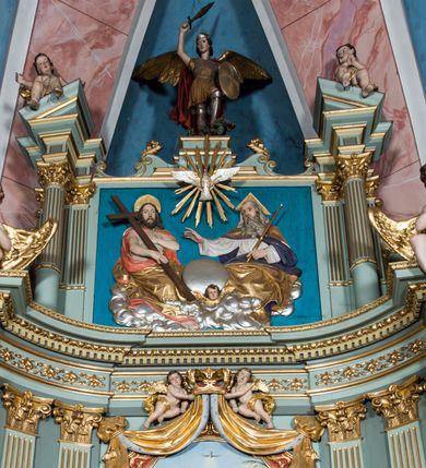 Zdjęcie nr 29: Ołtarz główny uzyskał formę przyściennej, dwukondygnacyjnej, neoklasycystycznej nastawy wzniesionej na rzucie elipsy z bocznymi bramkami. Stipes ołtarzowy uzyskał kształt leżącego prostopadłościanu podtrzymującego drewnianą mensę z portatylem. Jego lico dekorowane jest trzema równoległymi płycinami utworzonymi ze złoconych, profilowanych listew. Na mensie ustawiono prostopadłościenne tabernakulum w formie skrzynki flankowane parą pilastrów podtrzymujących nastawę tronu eucharystycznego. Ujmują go po bokach pary złoconych kolumn z korynckimi kapitelami podtrzymujące belkowanie wygięte na osi półkoliście. Na zwieńczeniu tabernakulum ustawiono leżącego na zamkniętej księdze z siedmioma pieczęciami srebrnego baranka apokaliptycznego na tle srebrzonych promieni. Pomiędzy kolumnami w polu zainstalowano obrotowy bęben z dwiema półkolistymi niszami. Retabulum zostało wsparte na wysokim cokole. Dolną kondygnację artykułują zwielokrotnione pilastry o kanelowanych trzonach i złoconych, kompozytowych głowicach oraz dwie wysunięte do przodu kanelowane kolumny o kapitelach kompozytowych. Podpory podtrzymują szeroki architraw wklęsło-wypukły dekorowany motywami roślinnymi oraz perełkami i ząbkami, wyłamany na przedłużeniu kolumn. W miejscu wyłamań usytuowano siedzących dwóch uskrzydlonych aniołów. Po bokach kolumn na konsolach ustawiono figury św. Piotra i św. Pawła. Boki ołtarza zamykają ażurowe wici roślinne pełniące rolę zredukowanych uszaków. W polu środkowym retabulum umieszczono prostokątne obramienie utworzone z dekoracyjnej ramy zdobionej wicią roślinną oraz podwieszonego u jej góry paludamentum z koroną podtrzymywaną przez dwa aniołki. W środku zainstalowano obraz Matki Boskiej Śnieżnej, a na zasuwie przedstawienie Chrztu Chrystusa w Jordanie. Zwieńczenie nastawy uzyskało kształt leżącego prostokąta flankowanego po bokach parą kanelowanych pilastrów o korynckich głowicach, na tle których ustawiono dwie kolumny podtrzymujących przerwane belkowanie z przerwanym przyczółkiem. Na osi zwieńczenia na postumencie ustawiono rzeźbę św. Michała Archanioła, a na przyczółkach rzeźby dwóch siedzących aniołów. W polu między pilastrami i kolumnami znalazło się płaskorzeźbione przedstawienie Trójcy Przenajświętszej. Dwie flankujące retabulum bramki ozdobiono dwoma aniołkami siedzącymi na ich zwieńczeniu na postumencie. Struktura całości pomalowane jest na zielono, ornamenty i profile wyzłocono, a rzeźby otrzymały naturalistyczną polichromię.