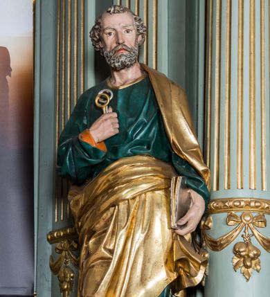Zdjęcie nr 1: Pełnoplastyczna rzeźba św. Piotra ukazuje stojącego apostoła frontalnie w nieznacznym kontrapoście. Święty prawą ręką przyciska do piersi dwa klucze – srebrny i złoty, a w lewej opuszczonej wzdłuż tułowia trzyma zamkniętą księgę. Piotr ma pociągłą twarz o ostrych rysach dojrzałego mężczyzny okoloną siwiejącą brodą i kędzierzawymi włosami. Charakteryzują ją wydatny nos i usta oraz spojrzenie szeroko otwartych oczu skierowane na widza. Apostoł ubrany jest w zieloną tunikę podbitą pomarańczem, lamowaną pod szyją złotą tasiemką oraz w złoty płaszcz narzucony na lewe ramię, z połą owinięta wokół pasa. Spod tuniki widać końcówki bosych stóp. 
