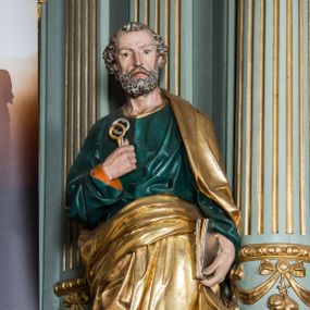 Zdjęcie nr 1: Pełnoplastyczna rzeźba św. Piotra ukazuje stojącego apostoła frontalnie w nieznacznym kontrapoście. Święty prawą ręką przyciska do piersi dwa klucze – srebrny i złoty, a w lewej opuszczonej wzdłuż tułowia trzyma zamkniętą księgę. Piotr ma pociągłą twarz o ostrych rysach dojrzałego mężczyzny okoloną siwiejącą brodą i kędzierzawymi włosami. Charakteryzują ją wydatny nos i usta oraz spojrzenie szeroko otwartych oczu skierowane na widza. Apostoł ubrany jest w zieloną tunikę podbitą pomarańczem, lamowaną pod szyją złotą tasiemką oraz w złoty płaszcz narzucony na lewe ramię, z połą owinięta wokół pasa. Spod tuniki widać końcówki bosych stóp. 