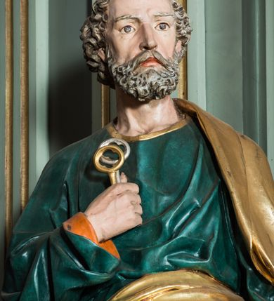 Zdjęcie nr 6: Pełnoplastyczna rzeźba św. Piotra ukazuje stojącego apostoła frontalnie w nieznacznym kontrapoście. Święty prawą ręką przyciska do piersi dwa klucze – srebrny i złoty, a w lewej opuszczonej wzdłuż tułowia trzyma zamkniętą księgę. Piotr ma pociągłą twarz o ostrych rysach dojrzałego mężczyzny okoloną siwiejącą brodą i kędzierzawymi włosami. Charakteryzują ją wydatny nos i usta oraz spojrzenie szeroko otwartych oczu skierowane na widza. Apostoł ubrany jest w zieloną tunikę podbitą pomarańczem, lamowaną pod szyją złotą tasiemką oraz w złoty płaszcz narzucony na lewe ramię, z połą owinięta wokół pasa. Spod tuniki widać końcówki bosych stóp. 