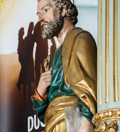 Zdjęcie nr 4: Pełnoplastyczna rzeźba św. Piotra ukazuje stojącego apostoła frontalnie w nieznacznym kontrapoście. Święty prawą ręką przyciska do piersi dwa klucze – srebrny i złoty, a w lewej opuszczonej wzdłuż tułowia trzyma zamkniętą księgę. Piotr ma pociągłą twarz o ostrych rysach dojrzałego mężczyzny okoloną siwiejącą brodą i kędzierzawymi włosami. Charakteryzują ją wydatny nos i usta oraz spojrzenie szeroko otwartych oczu skierowane na widza. Apostoł ubrany jest w zieloną tunikę podbitą pomarańczem, lamowaną pod szyją złotą tasiemką oraz w złoty płaszcz narzucony na lewe ramię, z połą owinięta wokół pasa. Spod tuniki widać końcówki bosych stóp. 