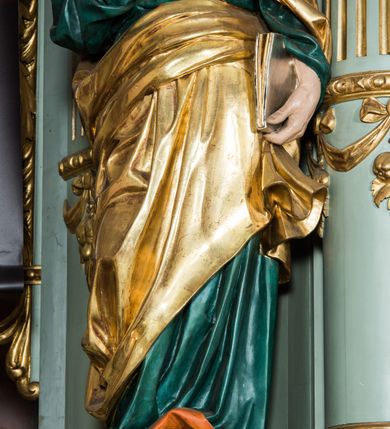 Zdjęcie nr 5: Pełnoplastyczna rzeźba św. Piotra ukazuje stojącego apostoła frontalnie w nieznacznym kontrapoście. Święty prawą ręką przyciska do piersi dwa klucze – srebrny i złoty, a w lewej opuszczonej wzdłuż tułowia trzyma zamkniętą księgę. Piotr ma pociągłą twarz o ostrych rysach dojrzałego mężczyzny okoloną siwiejącą brodą i kędzierzawymi włosami. Charakteryzują ją wydatny nos i usta oraz spojrzenie szeroko otwartych oczu skierowane na widza. Apostoł ubrany jest w zieloną tunikę podbitą pomarańczem, lamowaną pod szyją złotą tasiemką oraz w złoty płaszcz narzucony na lewe ramię, z połą owinięta wokół pasa. Spod tuniki widać końcówki bosych stóp. 