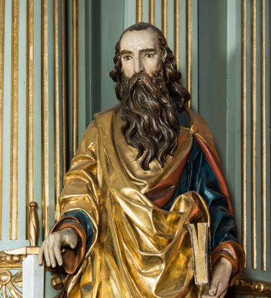 Zdjęcie nr 1: Pełnoplastyczna rzeźba św. Pawła ukazuje stojącego apostoła frontalnie w nieznacznym kontrapoście. Święty prawą rękę opiera na jelcu stojącego miecza, a w lewej opuszczonej wzdłuż tułowia trzyma zamkniętą księgę. Paweł ma pociągłą twarz o łysiejącym czole i ostrych rysach dojrzałego mężczyzny okoloną długą, mocno pofalowaną brodą i kędzierzawymi włosami. Charakteryzują ją wydatny nos i usta oraz spojrzenie szeroko otwartych oczu skierowane na widza. Apostoł ubrany jest w niebieską tunikę podbitą pomarańczem oraz w złoty płaszcz narzucony na ramiona i spięty na lewym barku z prawą połą owinięta wokół postaci. Płaszcz ma czerwone podbicie. Spod tuniki widać końcówki bosych stóp. 