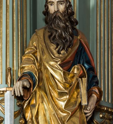 Zdjęcie nr 2: Pełnoplastyczna rzeźba św. Pawła ukazuje stojącego apostoła frontalnie w nieznacznym kontrapoście. Święty prawą rękę opiera na jelcu stojącego miecza, a w lewej opuszczonej wzdłuż tułowia trzyma zamkniętą księgę. Paweł ma pociągłą twarz o łysiejącym czole i ostrych rysach dojrzałego mężczyzny okoloną długą, mocno pofalowaną brodą i kędzierzawymi włosami. Charakteryzują ją wydatny nos i usta oraz spojrzenie szeroko otwartych oczu skierowane na widza. Apostoł ubrany jest w niebieską tunikę podbitą pomarańczem oraz w złoty płaszcz narzucony na ramiona i spięty na lewym barku z prawą połą owinięta wokół postaci. Płaszcz ma czerwone podbicie. Spod tuniki widać końcówki bosych stóp. 