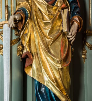 Zdjęcie nr 3: Pełnoplastyczna rzeźba św. Pawła ukazuje stojącego apostoła frontalnie w nieznacznym kontrapoście. Święty prawą rękę opiera na jelcu stojącego miecza, a w lewej opuszczonej wzdłuż tułowia trzyma zamkniętą księgę. Paweł ma pociągłą twarz o łysiejącym czole i ostrych rysach dojrzałego mężczyzny okoloną długą, mocno pofalowaną brodą i kędzierzawymi włosami. Charakteryzują ją wydatny nos i usta oraz spojrzenie szeroko otwartych oczu skierowane na widza. Apostoł ubrany jest w niebieską tunikę podbitą pomarańczem oraz w złoty płaszcz narzucony na ramiona i spięty na lewym barku z prawą połą owinięta wokół postaci. Płaszcz ma czerwone podbicie. Spod tuniki widać końcówki bosych stóp. 