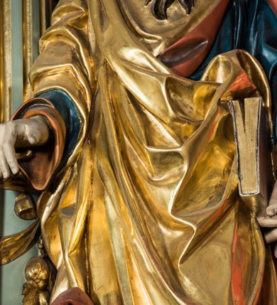 Zdjęcie nr 6: Pełnoplastyczna rzeźba św. Pawła ukazuje stojącego apostoła frontalnie w nieznacznym kontrapoście. Święty prawą rękę opiera na jelcu stojącego miecza, a w lewej opuszczonej wzdłuż tułowia trzyma zamkniętą księgę. Paweł ma pociągłą twarz o łysiejącym czole i ostrych rysach dojrzałego mężczyzny okoloną długą, mocno pofalowaną brodą i kędzierzawymi włosami. Charakteryzują ją wydatny nos i usta oraz spojrzenie szeroko otwartych oczu skierowane na widza. Apostoł ubrany jest w niebieską tunikę podbitą pomarańczem oraz w złoty płaszcz narzucony na ramiona i spięty na lewym barku z prawą połą owinięta wokół postaci. Płaszcz ma czerwone podbicie. Spod tuniki widać końcówki bosych stóp. 