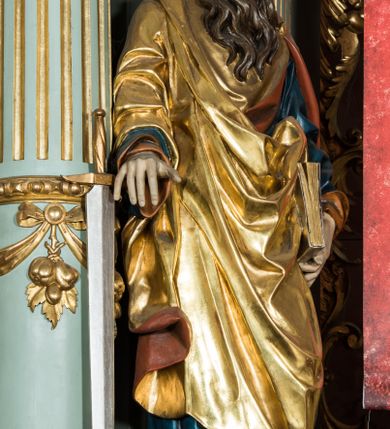 Zdjęcie nr 4: Pełnoplastyczna rzeźba św. Pawła ukazuje stojącego apostoła frontalnie w nieznacznym kontrapoście. Święty prawą rękę opiera na jelcu stojącego miecza, a w lewej opuszczonej wzdłuż tułowia trzyma zamkniętą księgę. Paweł ma pociągłą twarz o łysiejącym czole i ostrych rysach dojrzałego mężczyzny okoloną długą, mocno pofalowaną brodą i kędzierzawymi włosami. Charakteryzują ją wydatny nos i usta oraz spojrzenie szeroko otwartych oczu skierowane na widza. Apostoł ubrany jest w niebieską tunikę podbitą pomarańczem oraz w złoty płaszcz narzucony na ramiona i spięty na lewym barku z prawą połą owinięta wokół postaci. Płaszcz ma czerwone podbicie. Spod tuniki widać końcówki bosych stóp. 