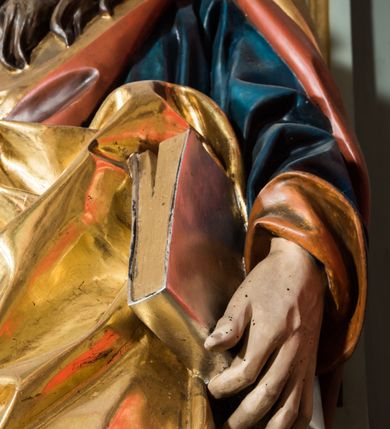 Zdjęcie nr 5: Pełnoplastyczna rzeźba św. Pawła ukazuje stojącego apostoła frontalnie w nieznacznym kontrapoście. Święty prawą rękę opiera na jelcu stojącego miecza, a w lewej opuszczonej wzdłuż tułowia trzyma zamkniętą księgę. Paweł ma pociągłą twarz o łysiejącym czole i ostrych rysach dojrzałego mężczyzny okoloną długą, mocno pofalowaną brodą i kędzierzawymi włosami. Charakteryzują ją wydatny nos i usta oraz spojrzenie szeroko otwartych oczu skierowane na widza. Apostoł ubrany jest w niebieską tunikę podbitą pomarańczem oraz w złoty płaszcz narzucony na ramiona i spięty na lewym barku z prawą połą owinięta wokół postaci. Płaszcz ma czerwone podbicie. Spod tuniki widać końcówki bosych stóp. 