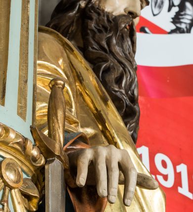 Zdjęcie nr 7: Pełnoplastyczna rzeźba św. Pawła ukazuje stojącego apostoła frontalnie w nieznacznym kontrapoście. Święty prawą rękę opiera na jelcu stojącego miecza, a w lewej opuszczonej wzdłuż tułowia trzyma zamkniętą księgę. Paweł ma pociągłą twarz o łysiejącym czole i ostrych rysach dojrzałego mężczyzny okoloną długą, mocno pofalowaną brodą i kędzierzawymi włosami. Charakteryzują ją wydatny nos i usta oraz spojrzenie szeroko otwartych oczu skierowane na widza. Apostoł ubrany jest w niebieską tunikę podbitą pomarańczem oraz w złoty płaszcz narzucony na ramiona i spięty na lewym barku z prawą połą owinięta wokół postaci. Płaszcz ma czerwone podbicie. Spod tuniki widać końcówki bosych stóp. 