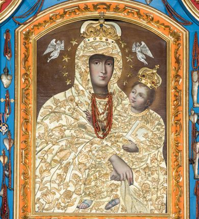 Zdjęcie nr 1: Obraz uzyskał kształt stojącego prostokąta. W centrum kompozycji przedstawiono frontalnie w półpostaci Matkę Boską z Dzieciątkiem na lewym ramieniu. Obie postacie przykrywają srebrne, złocone i trybowane w motywy kwiatowo-roślinne sukienki wotywne. Maria podtrzymuje małego Jezusa obiema, skrzyżowanymi rękami, trzymając równocześnie w prawej dłoni chustę. Jej głowa została ujęta nieznacznie w trzech czwartych w prawo. Madonna ma pociągłą twarz o jasnej karnacji i delikatnych, łagodnych rysach, z prostym nosem i pełnych ustach. Na głowie ma maforion lamowany taśmą. Maria ma na głowie złotą, zamkniętą koronę zwieńczoną globem z krzyżykiem. Korona jest dekorowaną  kameryzacją. Trzymane przez Madonnę Dzieciątko jest zwrócone w jej stronę w trzech czwartych. Mały Jezus lewą ręka podtrzymuje przy boku księgę Ewangelii. Natomiast nieznacznie usztywnioną i wyciągniętą przed siebie prawą rękę unosi w geście błogosławieństwa. Jezus ma okrągłą twarz o jasnej karnacji, pełnych ustach i rumianych policzkach, okoloną jasnobrązowymi, krótkimi włosami nad czołem układającymi się w loki. Ubrany jest w tunikę lamowaną taśmą pod szyją i przy rękawach. Na stopach widoczne są sandały. Na głowie ma zamknietą, kameryzowaną, złoconą i trybowaną w srebrze koronę wotywną. Głowy obu postaci otaczają świetliste nimby oraz dwanaście metalowych gwiazdek widocznych wokół głowy Matki Boskiej, a także dwóch srebrnych, trybowanych w blasze aniołów. Pod szyją Marii do sukienki przytwierdzono sznury korali oraz złote łańcuszki. u dołu bogato lamowaną złotym ornamentem roślinnym. 