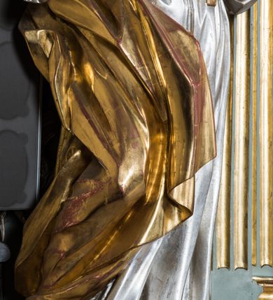 Zdjęcie nr 5: Pełnopostaciowa figura ukazuje stojącą frontalnie świętą męczenniczkę w kontrapoście. Święta z odchyloną nieznacznie do tyłu głową prawą ręką przyciska do piersi gałązkę palmową, lewą zaś na tej samej wysokości nieznacznie wyciąga do przodu. Kobieta ma pociągłą twarz o pucułowatych, rumianych policzkach, wyrazisty nos i usta. Jej głowę otaczają brązowe włosy widoczne spod złotej chusty. Ubrana jest w srebrzoną tunikę przewiązaną na piersi szarfą oraz narzucony na ramiona złoty płaszcz osuwający się z tyłu postaci. Spod tuniki widać fragment lewej stopy obutej w sandał.