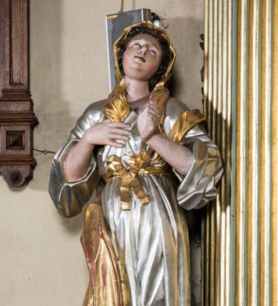 Zdjęcie nr 1: Pełnopostaciowa figura ukazuje stojącą frontalnie świętą męczenniczkę w kontrapoście. Święta z odchyloną nieznacznie do tyłu głową prawą ręką przyciska do piersi gałązkę palmową, lewą zaś na tej samej wysokości nieznacznie wyciąga do przodu. Kobieta ma pociągłą twarz o pucułowatych, rumianych policzkach, wyrazisty nos i usta. Jej głowę otaczają brązowe włosy widoczne spod złotej chusty. Ubrana jest w srebrzoną tunikę przewiązaną na piersi szarfą oraz narzucony na ramiona złoty płaszcz osuwający się z tyłu postaci. Spod tuniki widać fragment lewej stopy obutej w sandał.