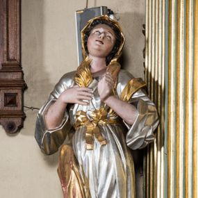 Zdjęcie nr 1: Pełnopostaciowa figura ukazuje stojącą frontalnie świętą męczenniczkę w kontrapoście. Święta z odchyloną nieznacznie do tyłu głową prawą ręką przyciska do piersi gałązkę palmową, lewą zaś na tej samej wysokości nieznacznie wyciąga do przodu. Kobieta ma pociągłą twarz o pucułowatych, rumianych policzkach, wyrazisty nos i usta. Jej głowę otaczają brązowe włosy widoczne spod złotej chusty. Ubrana jest w srebrzoną tunikę przewiązaną na piersi szarfą oraz narzucony na ramiona złoty płaszcz osuwający się z tyłu postaci. Spod tuniki widać fragment lewej stopy obutej w sandał.