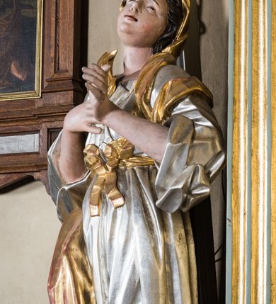 Zdjęcie nr 3: Pełnopostaciowa figura ukazuje stojącą frontalnie świętą męczenniczkę w kontrapoście. Święta z odchyloną nieznacznie do tyłu głową prawą ręką przyciska do piersi gałązkę palmową, lewą zaś na tej samej wysokości nieznacznie wyciąga do przodu. Kobieta ma pociągłą twarz o pucułowatych, rumianych policzkach, wyrazisty nos i usta. Jej głowę otaczają brązowe włosy widoczne spod złotej chusty. Ubrana jest w srebrzoną tunikę przewiązaną na piersi szarfą oraz narzucony na ramiona złoty płaszcz osuwający się z tyłu postaci. Spod tuniki widać fragment lewej stopy obutej w sandał.