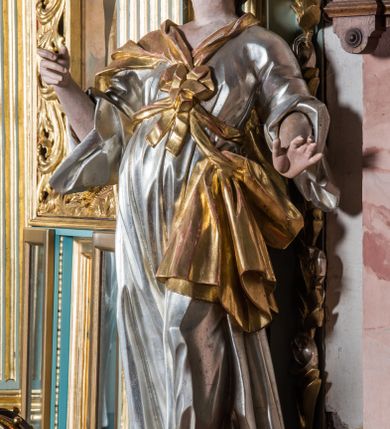 Zdjęcie nr 2: Pełnopostaciowa figura ukazuje stojącą frontalnie świętą niewiastę w kontrapoście. Święta z odchyloną nieznacznie do tyłu głową prawą ręką unosi na wysokości piersi trzymając w niej niezachowany przedmiot, lewą zaś lekko odsuwa od siebie. Kobieta ma pociągłą twarz o pucułowatych, rumianych policzkach, wyrazisty nos i usta. Jej głowę otaczają brązowe włosy spływające na ramiona. Ubrana jest w srebrzoną tunikę przewiązaną na piersi szarfą oraz narzucony na ramiona złoty płaszcz osuwający się z tyłu postaci. Spod tuniki widać fragment lewej stopy obutej w sandał.