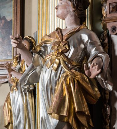 Zdjęcie nr 4: Pełnopostaciowa figura ukazuje stojącą frontalnie świętą niewiastę w kontrapoście. Święta z odchyloną nieznacznie do tyłu głową prawą ręką unosi na wysokości piersi trzymając w niej niezachowany przedmiot, lewą zaś lekko odsuwa od siebie. Kobieta ma pociągłą twarz o pucułowatych, rumianych policzkach, wyrazisty nos i usta. Jej głowę otaczają brązowe włosy spływające na ramiona. Ubrana jest w srebrzoną tunikę przewiązaną na piersi szarfą oraz narzucony na ramiona złoty płaszcz osuwający się z tyłu postaci. Spod tuniki widać fragment lewej stopy obutej w sandał.